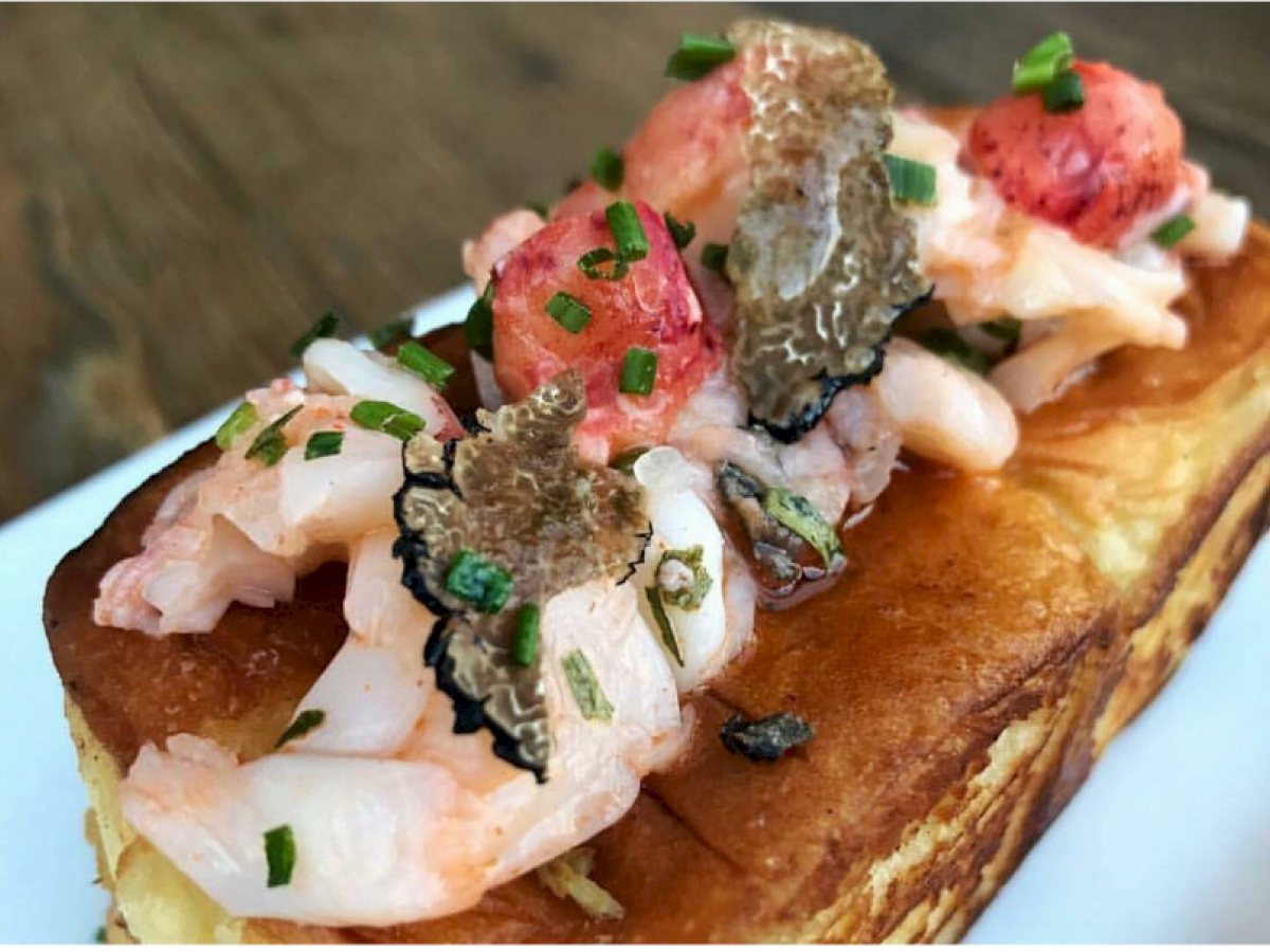 A gourmet toast topped with shrimp, lobster, black truffle slices, and chopped chives on a crispy bread slice, served on a white plate.