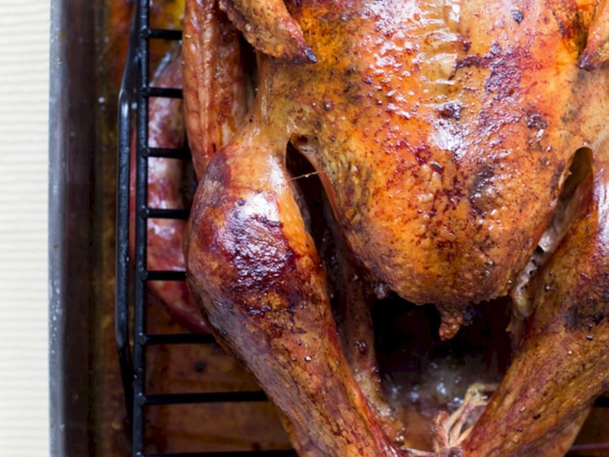 A roasted turkey sits in a roasting pan with a rack. The turkey has a golden-brown, crispy skin and is positioned breast-side up in the pan.