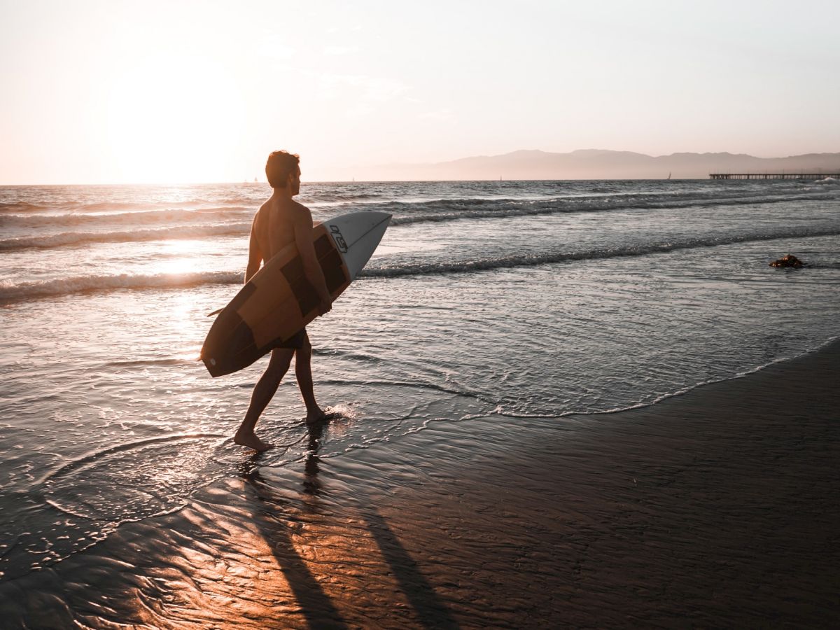 A person carrying a surfboard walks along the beach at sunset, with waves gently lapping at their feet and mountains visible in the background.