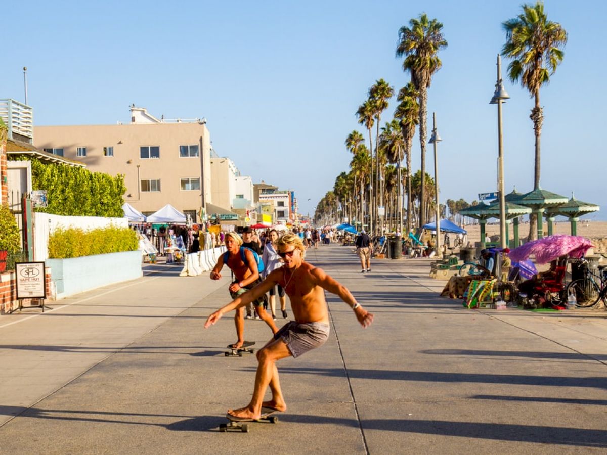 Two people are skateboarding on a sunny boardwalk, with palm trees, buildings, and stalls in the background.