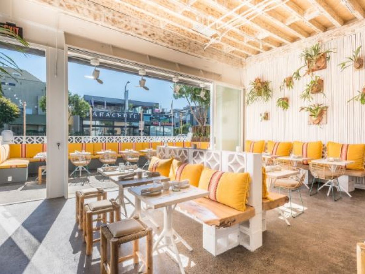 A bright, modern café with yellow cushions, wooden tables, and wall plants. Open-air design with a view of the street outside completes the cozy space.