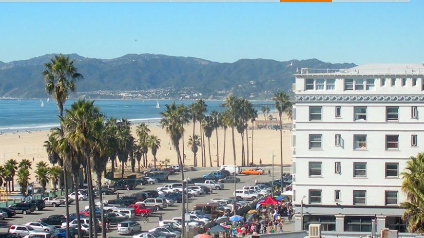 This image shows a beachfront scene with a large white building, palm trees, parked cars, and mountains in the background, with clear blue skies.
