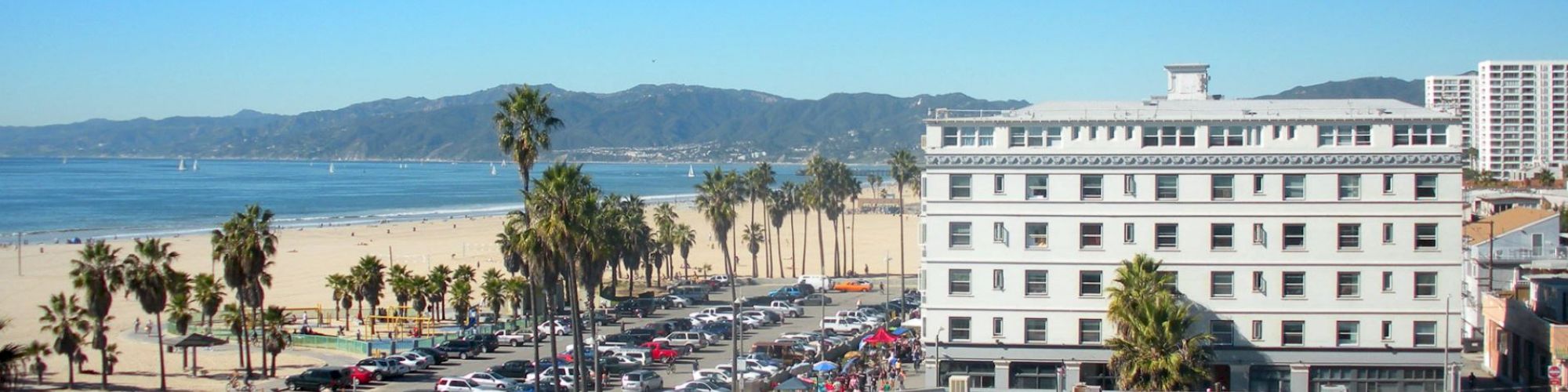 This image shows a beachfront scene with a large white building, palm trees, parked cars, and mountains in the background, with clear blue skies.