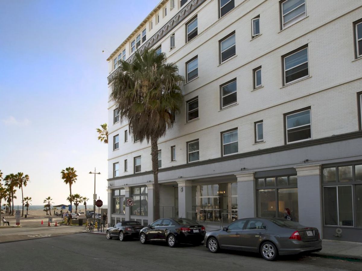 A street view of a multi-story building with parked cars, palm trees, and the ocean visible in the background under a clear sky.