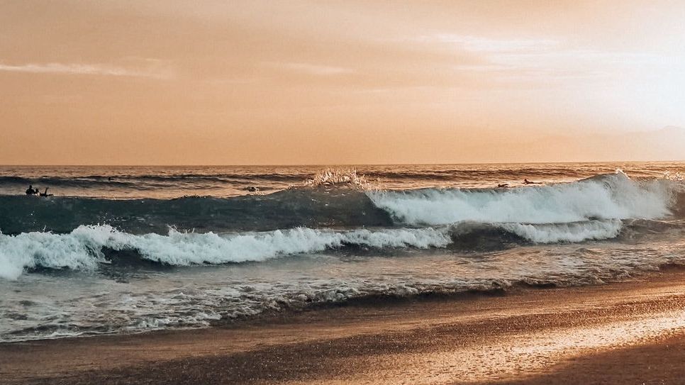 A serene beach scene during sunset with waves crashing against the shore and the sun casting a warm glow over the horizon.