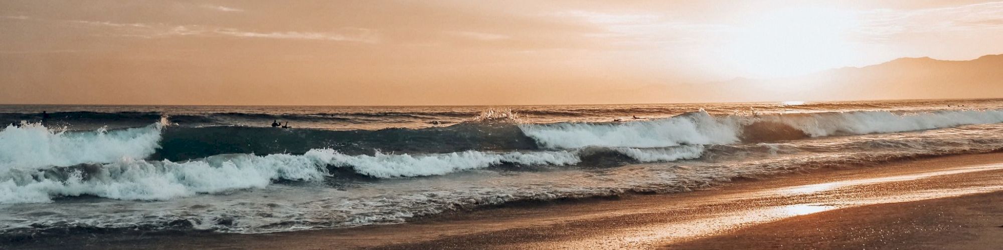 A serene beach scene during sunset with waves crashing against the shore and the sun casting a warm glow over the horizon.