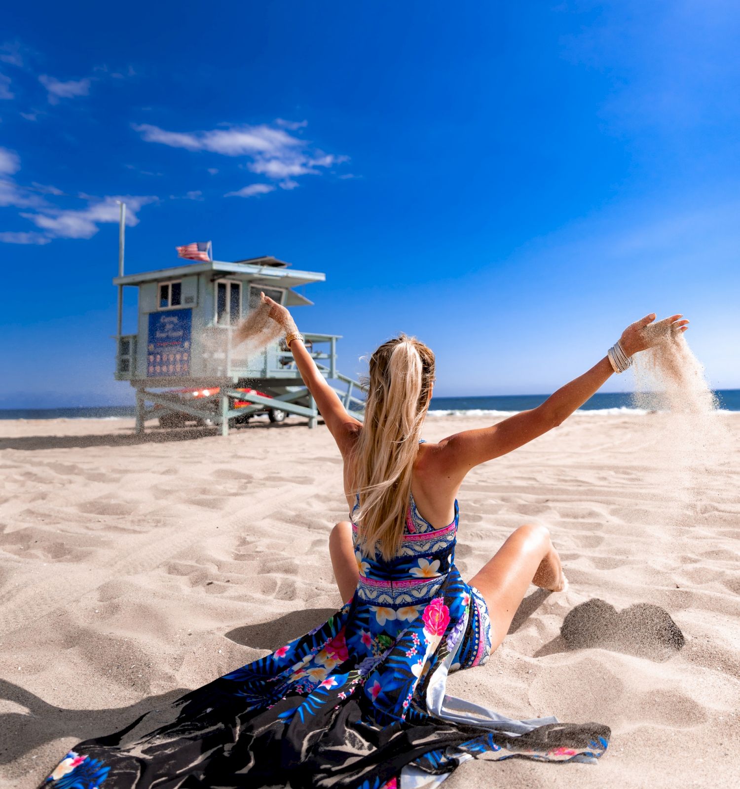 A person in a colorful dress sits on a sandy beach, arms raised, with a lifeguard tower and a clear blue sky in the background.