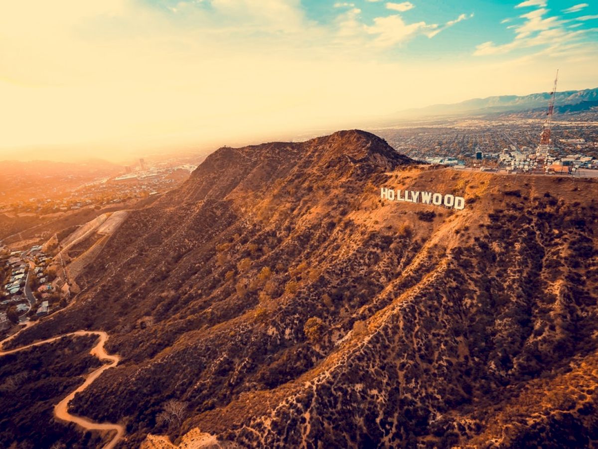 This image shows an aerial view of the famous Hollywood sign on a hilly landscape with a cityscape in the background under a dramatic sunset sky.