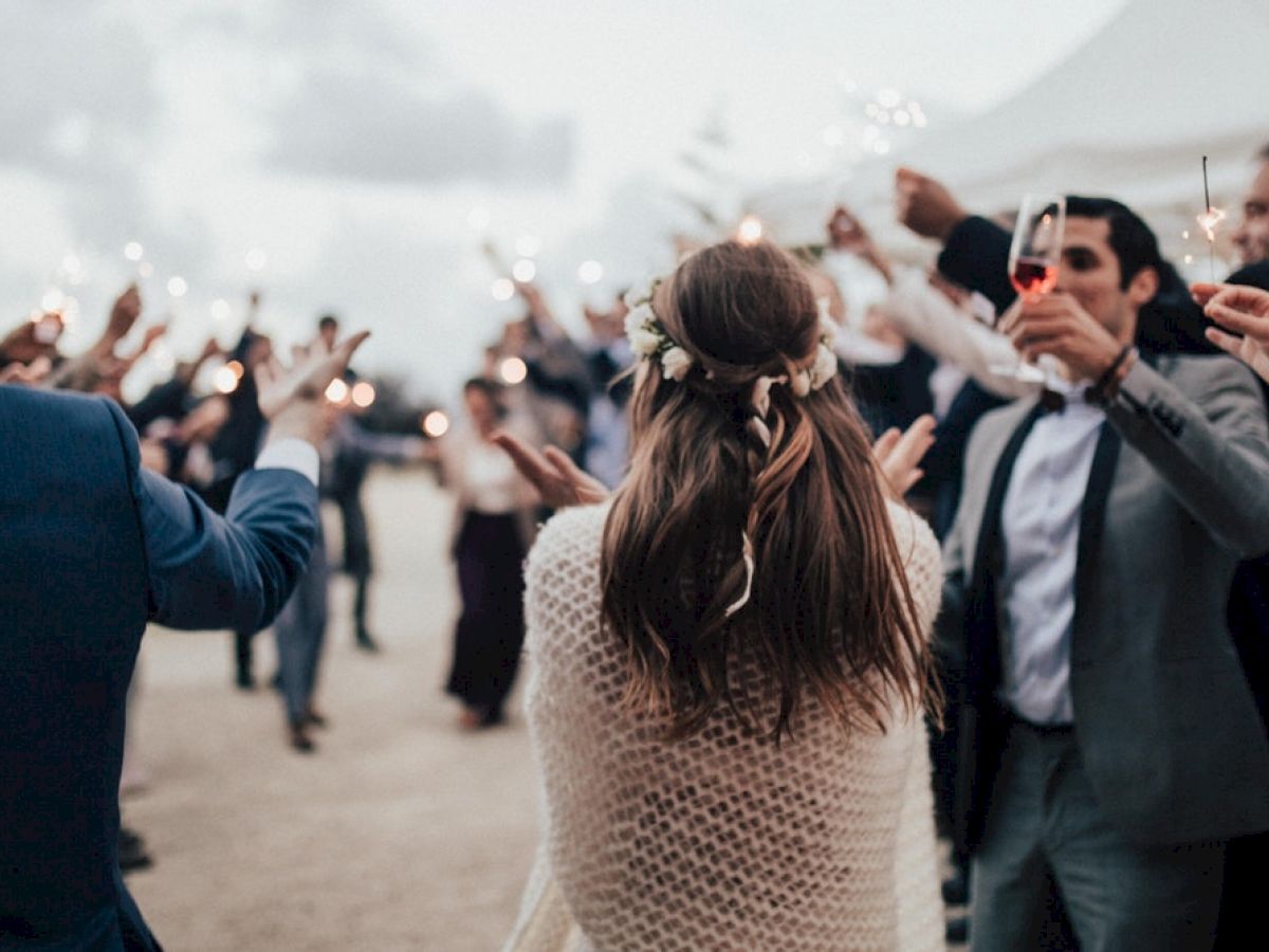 A group of people celebrate with sparklers outdoors, gathered around a person with long hair adorned with flowers, wearing a white shawl.