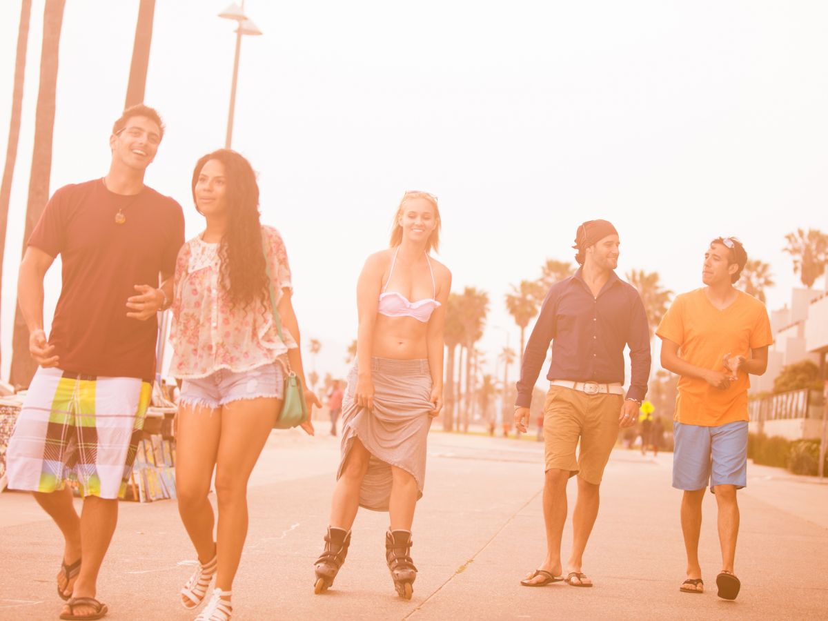 A group of five people walking on a sunny boardwalk, with palm trees in the background. One person is on rollerblades, and all are dressed casually.