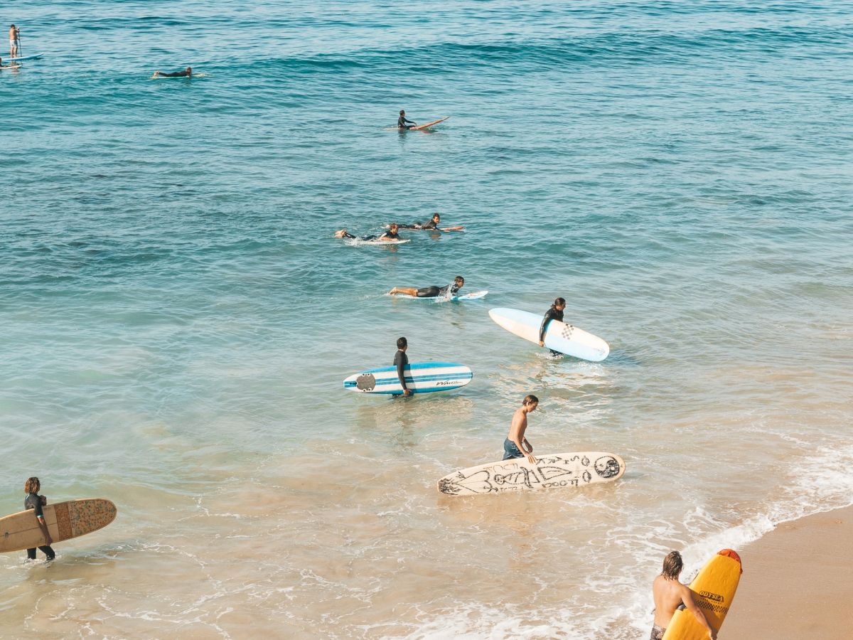 The image shows several surfers with their surfboards in the ocean and on a sandy beach, under a clear sky, enjoying a day by the water.