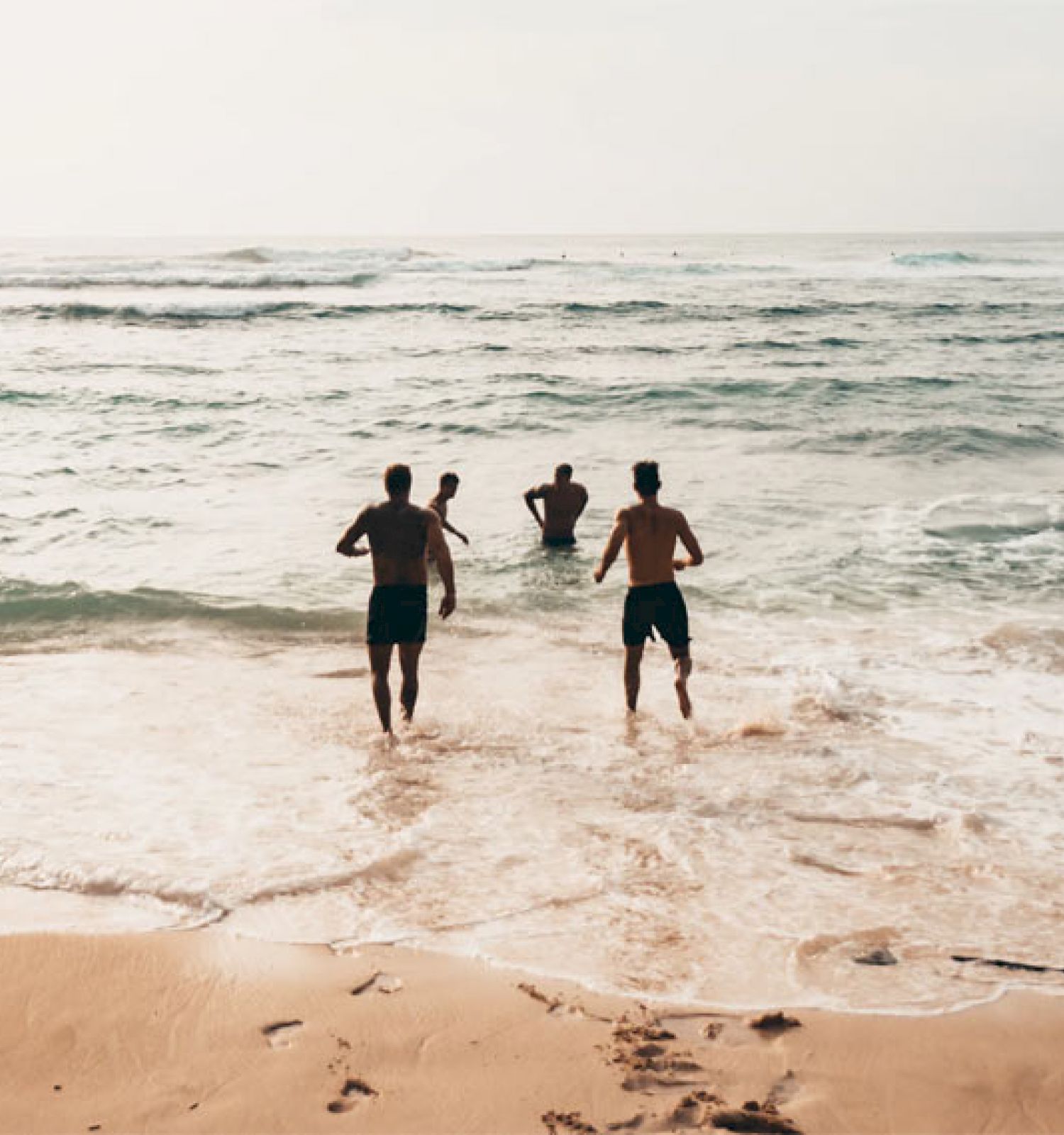 The image shows four people running into the waves at a beach, leaving footprints in the sand behind them.