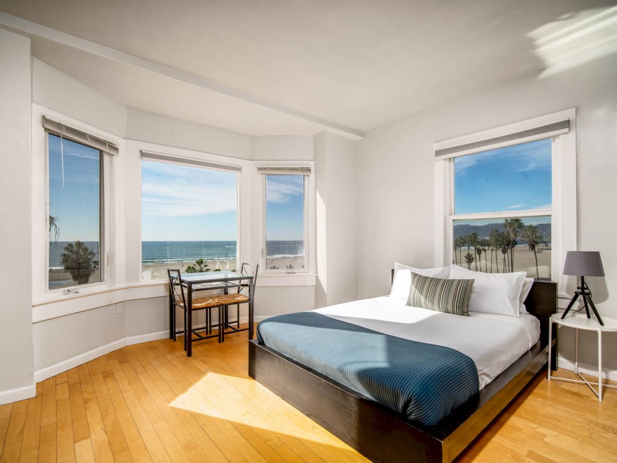 A bright bedroom with a double bed, nightstand, lamp, and a small desk by bay windows offering a scenic ocean view.