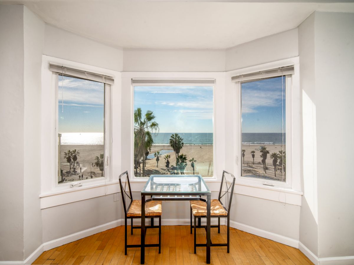 A small dining table with two chairs is set in a bay window, offering an oceanfront view with palm trees under a sunny sky.