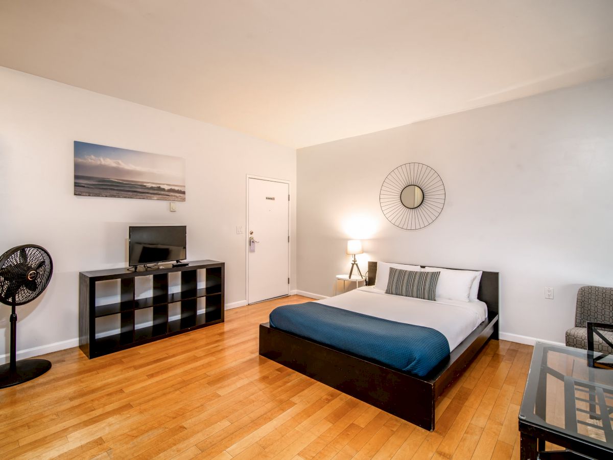 A minimalist bedroom with a bed, TV stand, fan, wall art, a lamp, and a mirror on the wall, featuring wooden flooring and modern decor.