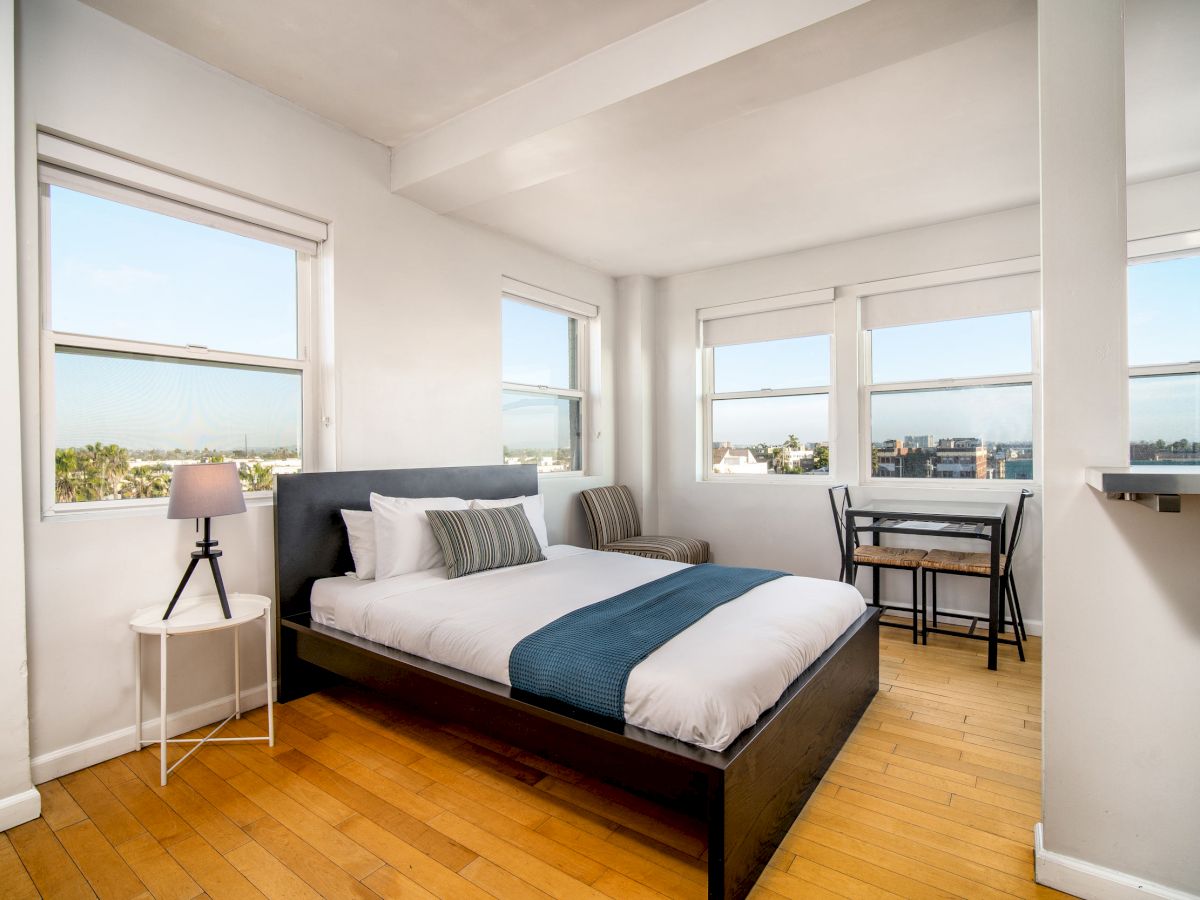 A bright bedroom with a double bed, blue accents, wooden flooring, three windows, a small side table with a lamp, and a desk with a chair.