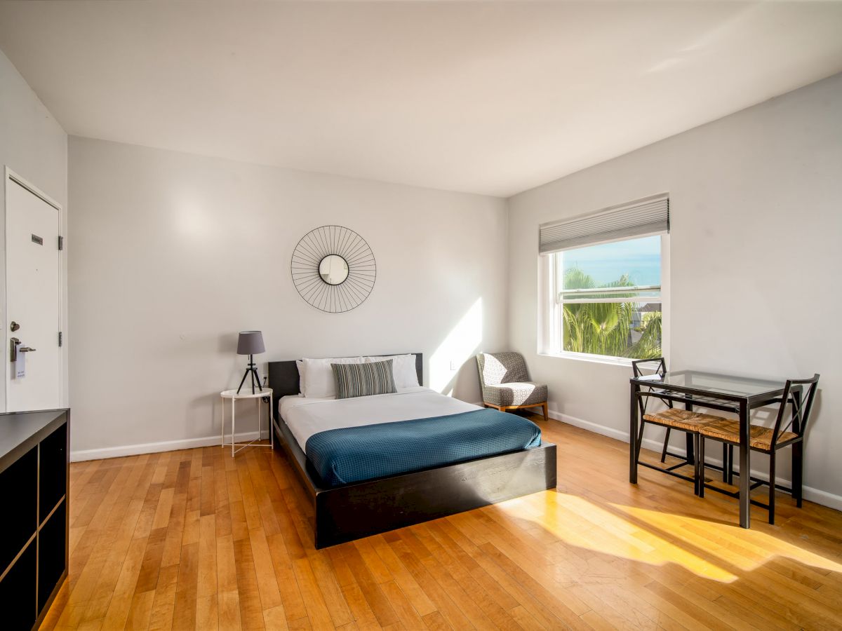 A minimalist bedroom with a bed, side table, chair, small dining set, wall decor, and a window allowing natural light.