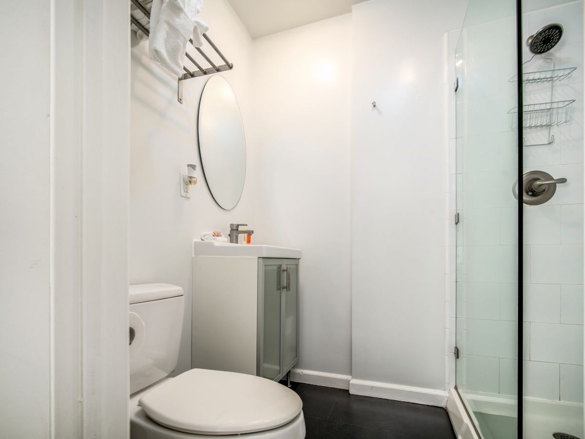 A small bathroom with a toilet, vanity with an oval mirror, and a glass-enclosed shower. A towel rack and shower caddy are also visible in the room.