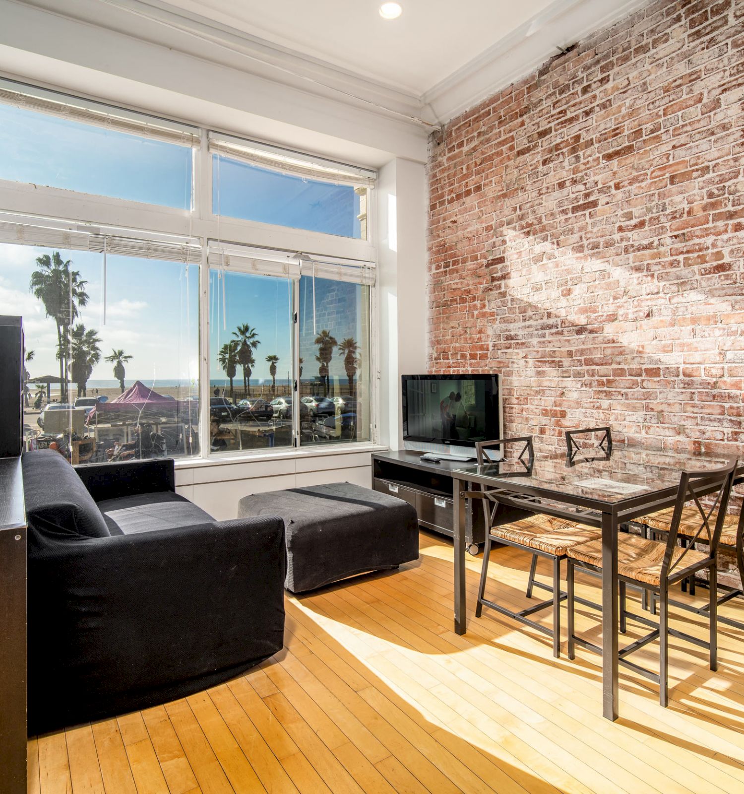 A modern living room with brick walls, large windows showing a sunny view, a black sofa, a TV, and a dining table with chairs.