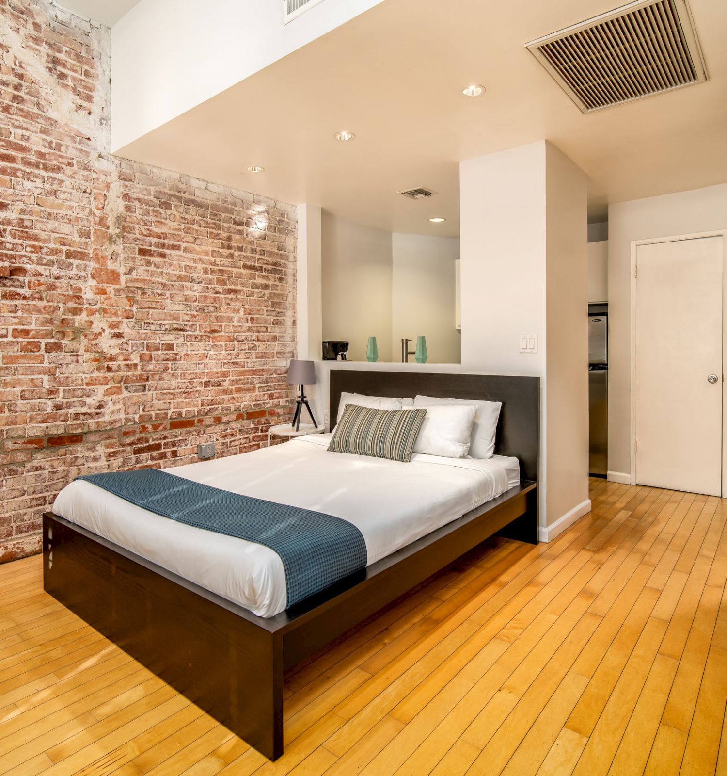 A modern bedroom with a brick accent wall, hardwood floors, a bed, a small table and chairs, and a door leading to an adjacent room ending the sentence.