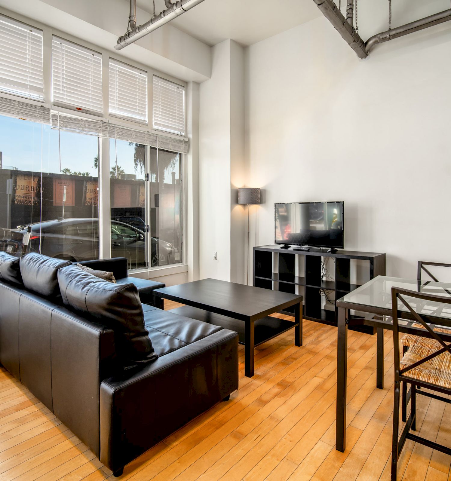 A modern living room with large windows, a black sofa, a TV on a stand, a glass dining table with chairs, and wooden floors.