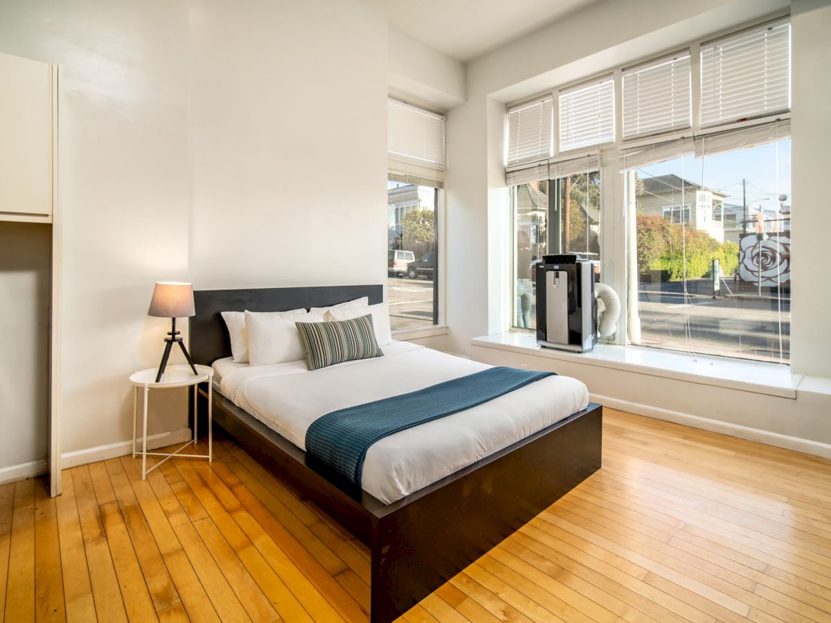 A minimalistic bedroom with a wooden floor, a bed with blue accents, a lamp on a side table, and large windows overlooking the street outside.