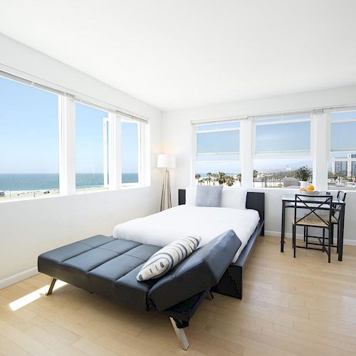 A minimalist bedroom with a large bed, black sofa, small table and chairs by the windows, and an ocean view through multiple windows.