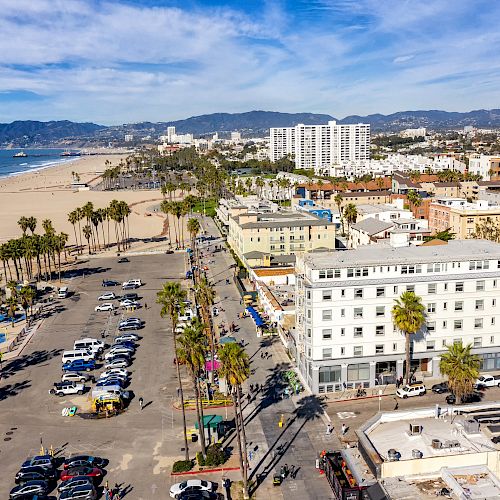 Aerial view of a coastal city with a beachfront, parking lot, palm trees, and surrounding buildings under a blue sky with scattered clouds.