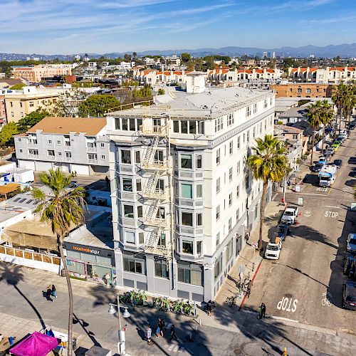 This image shows an aerial view of a cityscape featuring a multi-story building at a street intersection with palm trees and buildings.