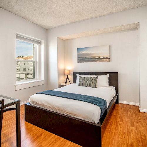 A tidy bedroom with a large bed, wooden floor, desk with chair, window, artwork on the wall, and a standing lamp in the corner ending the sentence.