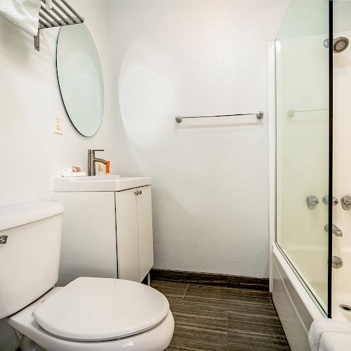 A small, modern bathroom with a toilet, vanity with a round mirror, towel rack, and a bathtub with a glass door and overhead shower.