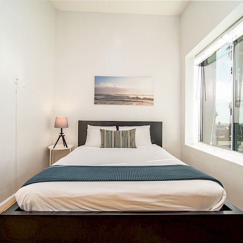 A minimalist bedroom with a neatly made bed, a side table with a lamp, and a large window with a view of palm trees and the ocean.