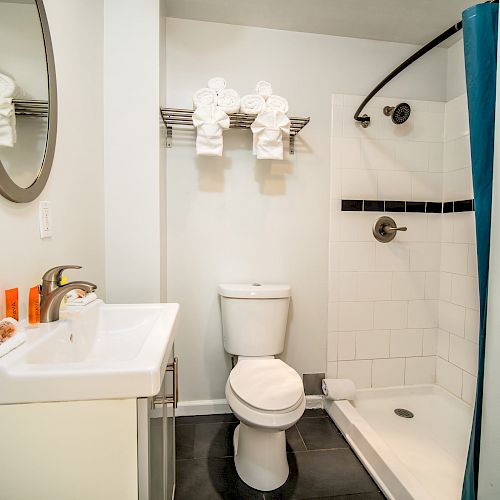 A small bathroom with a sink, mirror, toilet, shower, blue shower curtain, towel rack with towels, and toiletries on the sink.