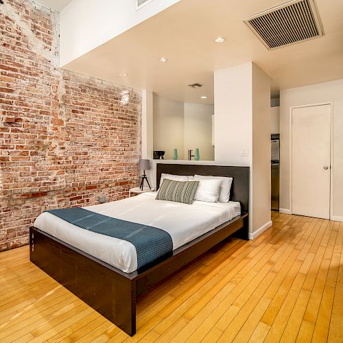 A modern bedroom with a brick accent wall, wooden floor, bed with white and blue linens, small dining area, and a white closet and partition wall.