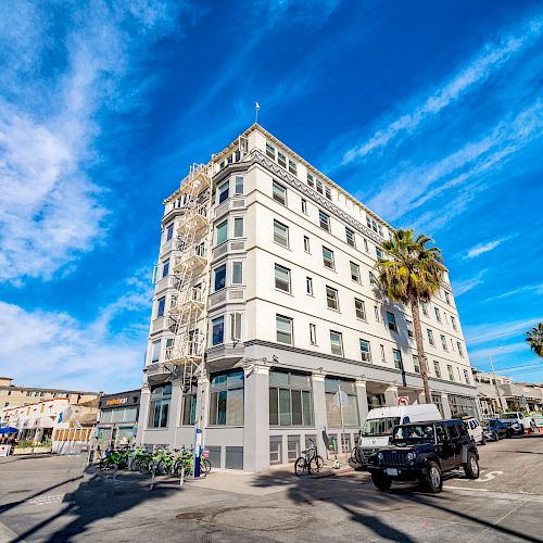 A multi-story building on a sunny day, surrounded by palm trees and parked vehicles, with a vibrant blue sky in the background and some clouds visible.