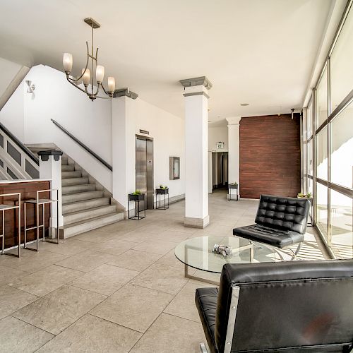 A modern lobby with seating, a table, a staircase, and an elevator; a bar area and large windows providing natural light.