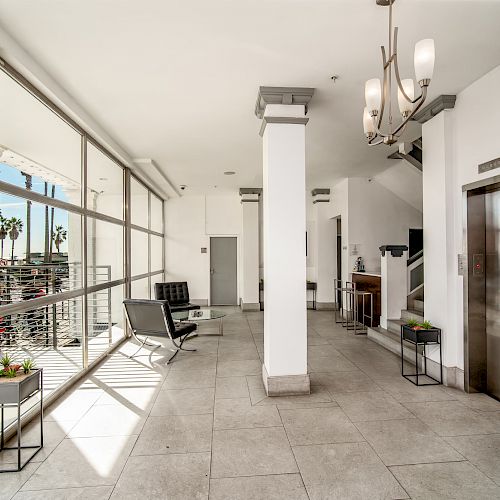 The image shows a modern lobby with floor-to-ceiling windows, seating, potted plants, an elevator, and a light fixture.