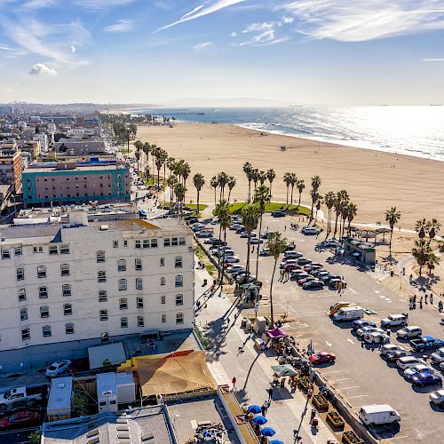 An aerial view of a beach with a parking lot, palm trees, buildings, and the ocean on a clear day.