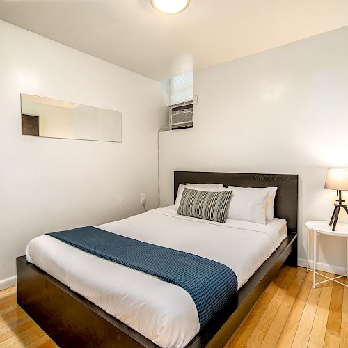 A minimalist bedroom features a double bed with white linens and a blue throw, a bedside table with a lamp, a wall mirror, and wooden flooring.