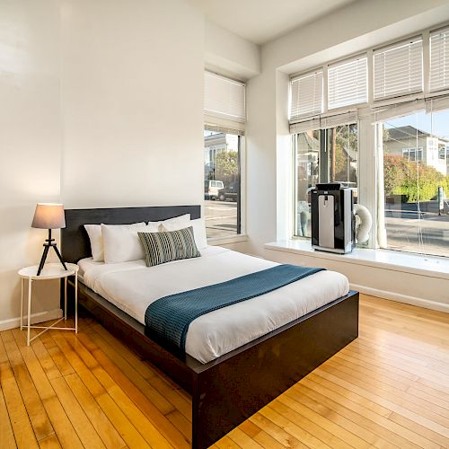 A minimalist bedroom with a large window, wooden floors, a bed with blue accents, a bedside table, and a lamp.