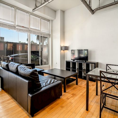 A modern living room features a black couch, coffee table, TV, and dining table with chairs, against large windows.