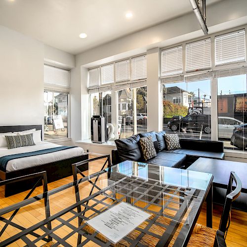 The image showcases a modern studio apartment with a large bed, glass dining table, and a black sectional sofa, featuring large windows and hardwood floors.