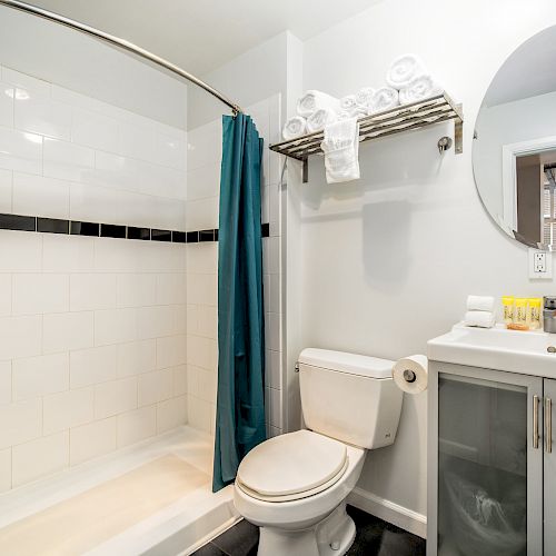 A bathroom with a shower, teal curtain, toilet, sink, round mirror, and shelves with towels and toiletries is shown ending the sentence.