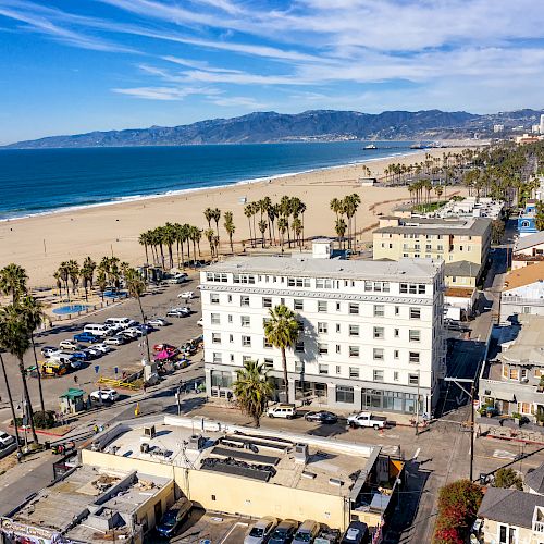 A beach with palm trees, a clear blue sky, buildings, cars, and mountains in the background.