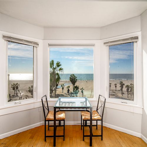 A small dining table with two chairs is set in front of a bay window, offering a panoramic view of a beach with palm trees and ocean.