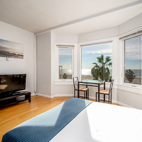 A bright room with a TV, a bed, a table with chairs by large windows showcasing a sea view and palm trees on a sunny day.