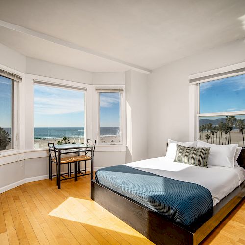 The image shows a bright bedroom with ocean view through large windows, featuring a double bed, a small desk and chair, and a nightstand with a lamp.