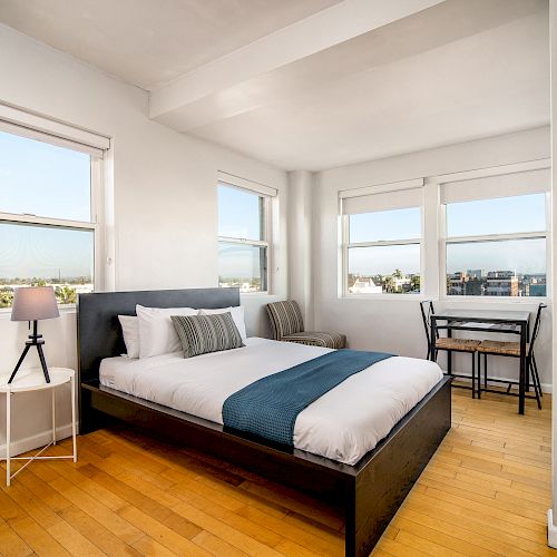 A minimalist bedroom with a bed, nightstand and lamp, three large windows, a small table with chairs, and hardwood floors, bright and airy.