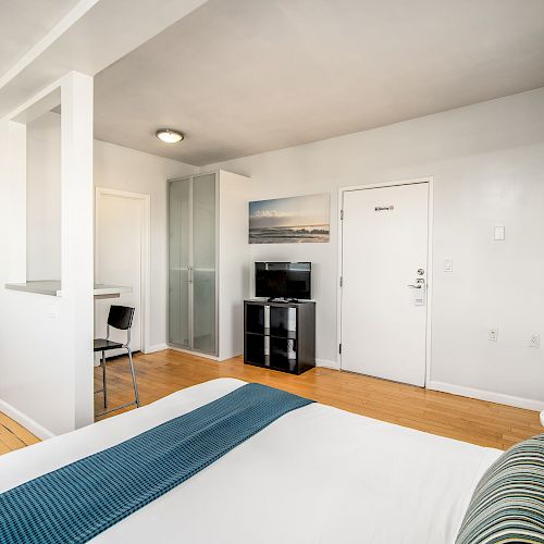 A modern, minimalist hotel room with a bed, TV, small table, and chair, featuring a clean design with white walls and a light wooden floor.