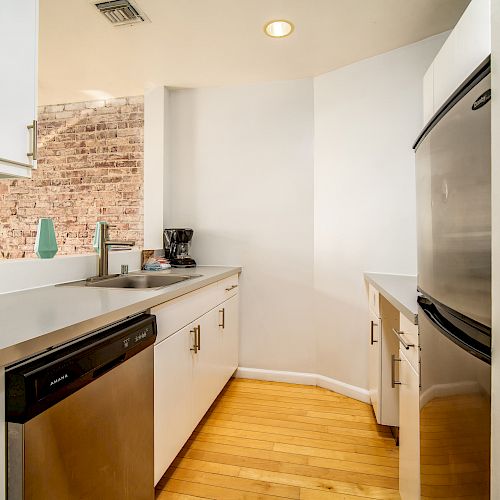 A modern kitchen with stainless steel appliances, white cabinetry, and a wooden floor. There is a sink, dishwasher, and coffee maker on the counter.